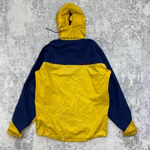 May include: A yellow and navy blue hooded jacket featuring a color-blocked design, a drawstring hood, long sleeves with adjustable cuffs, and the Mammut brand name printed in small red letters on the upper back panel of the garment.