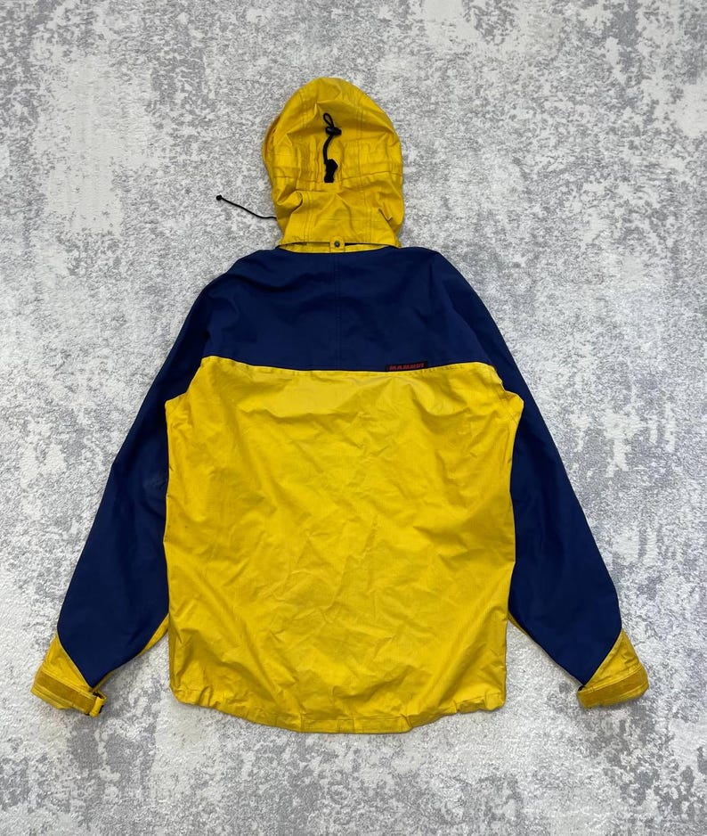 May include: A yellow and navy blue hooded jacket featuring a color-blocked design, a drawstring hood, long sleeves with adjustable cuffs, and the Mammut brand name printed in small red letters on the upper back panel of the garment.