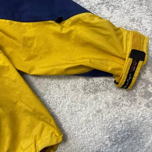 May include: A long-sleeve jacket featuring a vibrant yellow main body with dark blue panels on the upper shoulder area. The sleeve cuff includes a black adjustable hook-and-loop strap with the text Mammut and Gore-Tex printed in white.