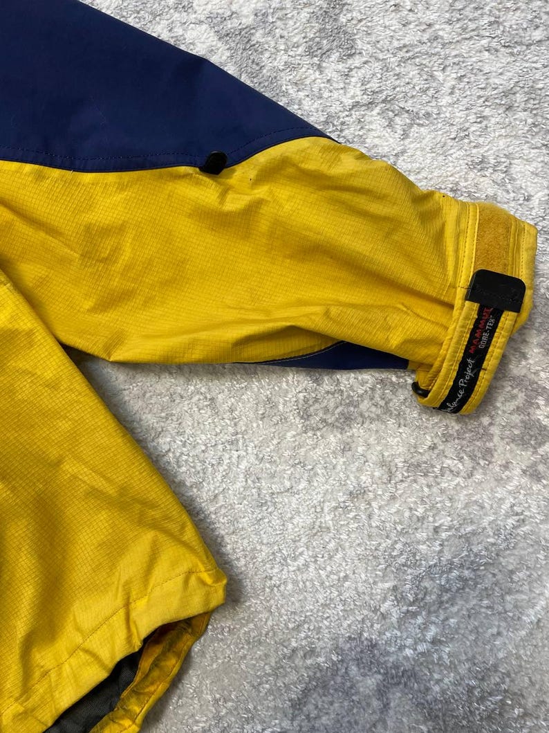 May include: A long-sleeve jacket featuring a vibrant yellow main body with dark blue panels on the upper shoulder area. The sleeve cuff includes a black adjustable hook-and-loop strap with the text Mammut and Gore-Tex printed in white.