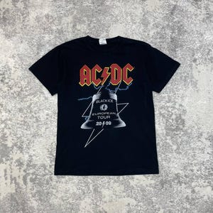Può includere: Una t-shirt nera a maniche corte con il logo AC/DC in rosso e giallo nella parte superiore. Sotto il logo è presente la grafica di una campana con il testo "BLACK ICE EUROPEAN TOUR 2009" e un fulmine bianco sul davanti della maglietta.