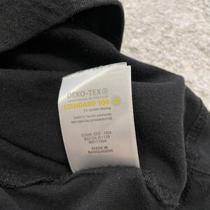 May include: Close-up of a black garment with a white label. The label reads "OEKO-TEX STANDARD 100" and includes text about testing for harmful substances. The label also includes a code, batch number, and the phrase "MADE IN BANGLADESH."