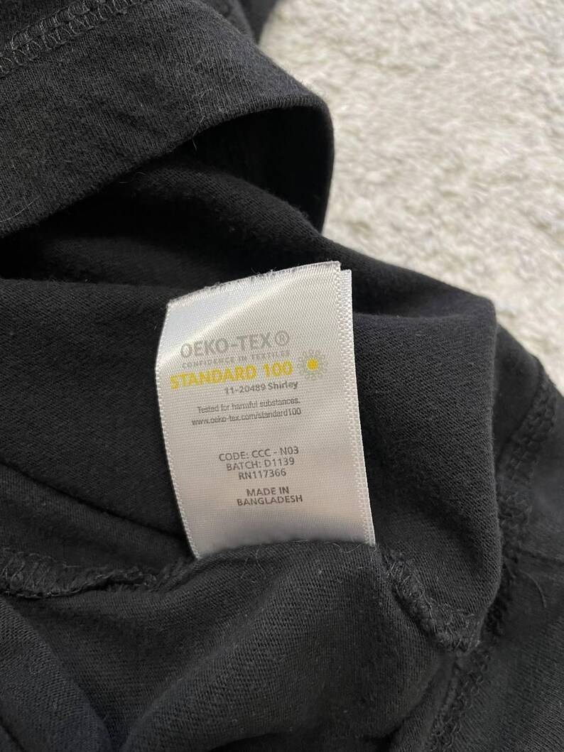 May include: Close-up of a black garment with a white label. The label reads "OEKO-TEX STANDARD 100" and includes text about testing for harmful substances. The label also includes a code, batch number, and the phrase "MADE IN BANGLADESH."