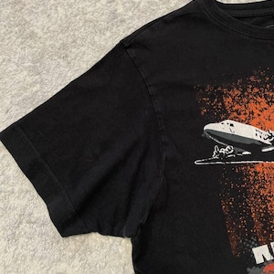 May include: Black t-shirt featuring a graphic of a zeppelin with orange and white accents. The shirt has a crew neck and short sleeves. The graphic is on the lower right side of the shirt.