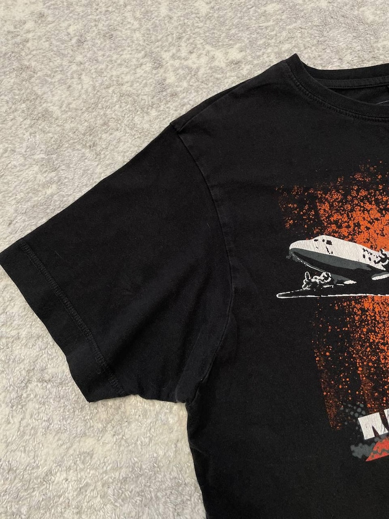 May include: Black t-shirt featuring a graphic of a zeppelin with orange and white accents. The shirt has a crew neck and short sleeves. The graphic is on the lower right side of the shirt.