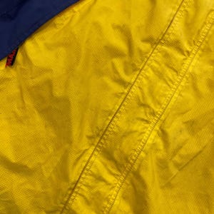 May include: A bright yellow jacket with a grid-like ripstop texture and a dark blue panel at the top. A small black tag with red text reading MAM is visible on the left side, attached to a seam. The garment features reinforced stitching.