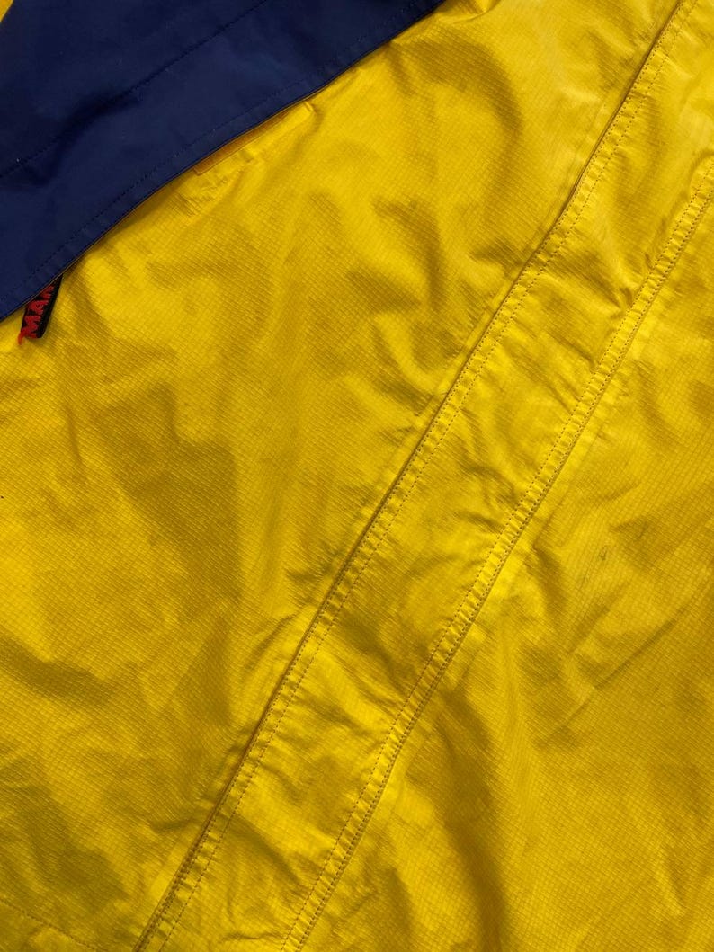 May include: A bright yellow jacket with a grid-like ripstop texture and a dark blue panel at the top. A small black tag with red text reading MAM is visible on the left side, attached to a seam. The garment features reinforced stitching.