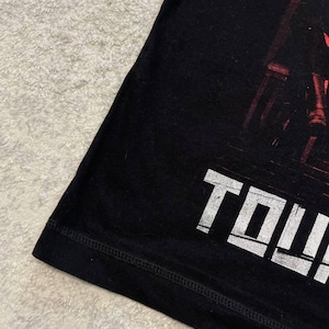 K&ouml;nnte beinhalten: Ein schwarzes T-Shirt mit einem Grafikdruck mit roten Akzenten und dem wei&szlig;en Text "TOW". Das Kleidungsst&uuml;ck hat einen sichtbaren Saum an der Unterkante und eine glatte Stoffstruktur, die einen Teil des Designs auf der Vorderseite zeigt.