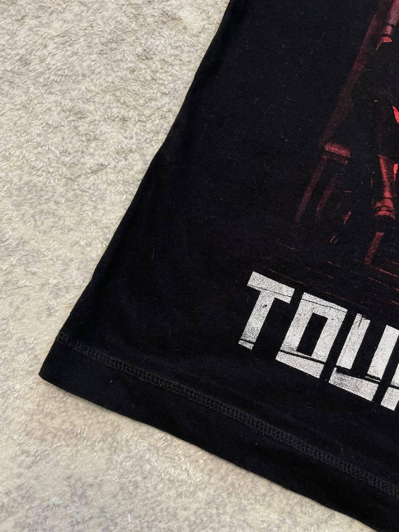 K&ouml;nnte beinhalten: Ein schwarzes T-Shirt mit einem Grafikdruck mit roten Akzenten und dem wei&szlig;en Text "TOW". Das Kleidungsst&uuml;ck hat einen sichtbaren Saum an der Unterkante und eine glatte Stoffstruktur, die einen Teil des Designs auf der Vorderseite zeigt.