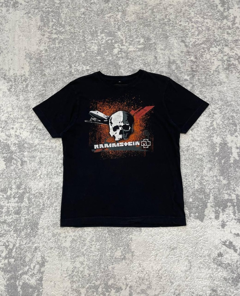 May include: Black t-shirt featuring a graphic design with a skull, a zeppelin, and the band name "Rammstein". The design incorporates red, orange, and white elements. The shirt is a classic crew neck style.
