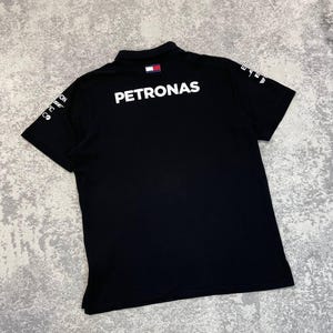 May include: Black polo shirt with the word "PETRONAS" in large white letters across the back. The shirt has a small logo on the collar and additional white text on the sleeves. The shirt is made of a soft material.