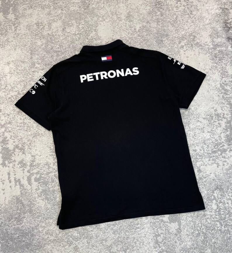 May include: Black polo shirt with the word "PETRONAS" in large white letters across the back. The shirt has a small logo on the collar and additional white text on the sleeves. The shirt is made of a soft material.