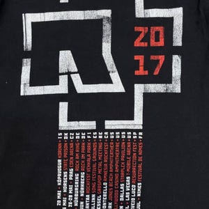 K&ouml;nnte beinhalten: Ein schwarzes T-Shirt mit einem gro&szlig;en wei&szlig;en kreuzartigen Logo und dem roten Text 2017. Unter dem Logo befindet sich eine Liste von Tourdaten und Orten in wei&szlig;em und rotem Text, darunter St&auml;dte wie Chicago, Dallas und Las Vegas auf dem Stoff.