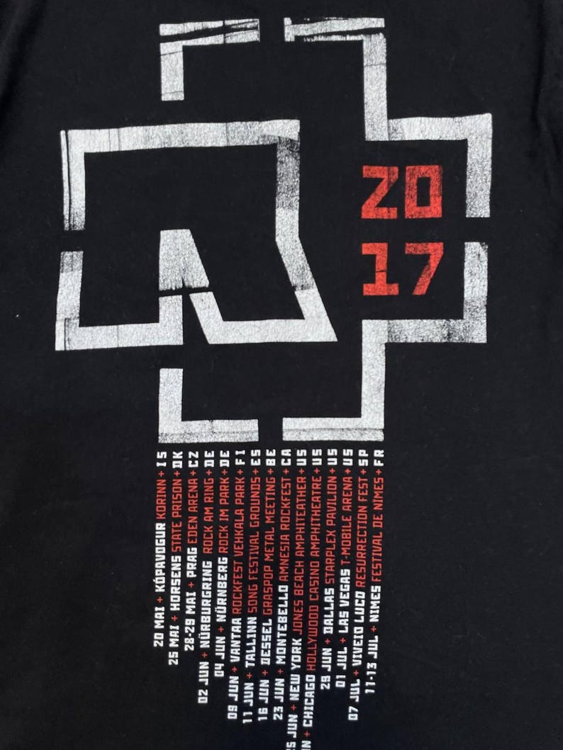 K&ouml;nnte beinhalten: Ein schwarzes T-Shirt mit einem gro&szlig;en wei&szlig;en kreuzartigen Logo und dem roten Text 2017. Unter dem Logo befindet sich eine Liste von Tourdaten und Orten in wei&szlig;em und rotem Text, darunter St&auml;dte wie Chicago, Dallas und Las Vegas auf dem Stoff.