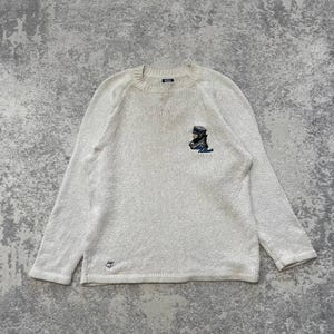 May include: Cream-coloured long-sleeved jumper with a crew neck. The jumper has a small embroidered design on the chest of a figure wearing a hat and coat. The jumper has a textured knit and a small logo near the bottom hem.