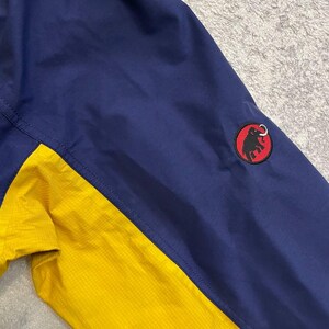 May include: A two-toned jacket featuring a dark blue upper section and a bright yellow lower section. A circular patch is stitched onto the dark blue sleeve, displaying a black silhouette of a mammoth on a red background with a black border.