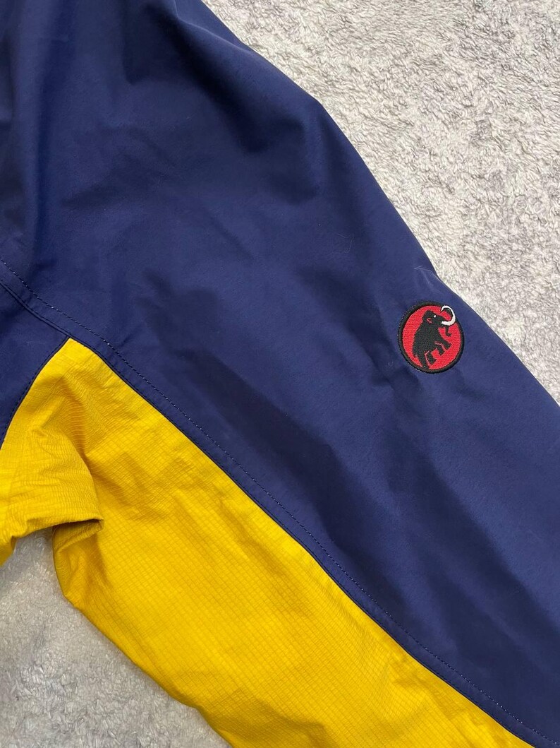 May include: A two-toned jacket featuring a dark blue upper section and a bright yellow lower section. A circular patch is stitched onto the dark blue sleeve, displaying a black silhouette of a mammoth on a red background with a black border.