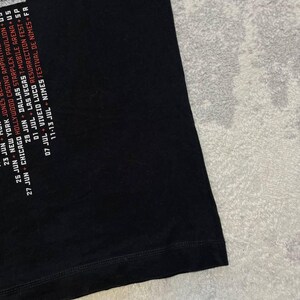 K&ouml;nnte beinhalten: Ein schwarzes T-Shirt mit wei&szlig;em und rotem Textdruck, der Konzerttourneedaten und Orte wie New York, Chicago, Dallas, Las Vegas, Viveiro und N&icirc;mes auflistet. Die Grafik befindet sich auf der linken Seite des schwarzen Stoffes.