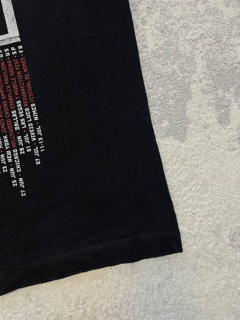 K&ouml;nnte beinhalten: Ein schwarzes T-Shirt mit wei&szlig;em und rotem Textdruck, der Konzerttourneedaten und Orte wie New York, Chicago, Dallas, Las Vegas, Viveiro und N&icirc;mes auflistet. Die Grafik befindet sich auf der linken Seite des schwarzen Stoffes.