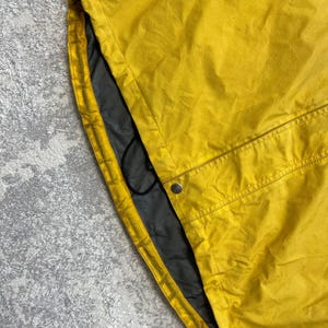 May include: A yellow jacket with a grid-like texture, featuring a dark blue panel at the top, a grey interior lining, a black drawstring cord, and a single silver-toned metal snap button closure near the bottom hem area.