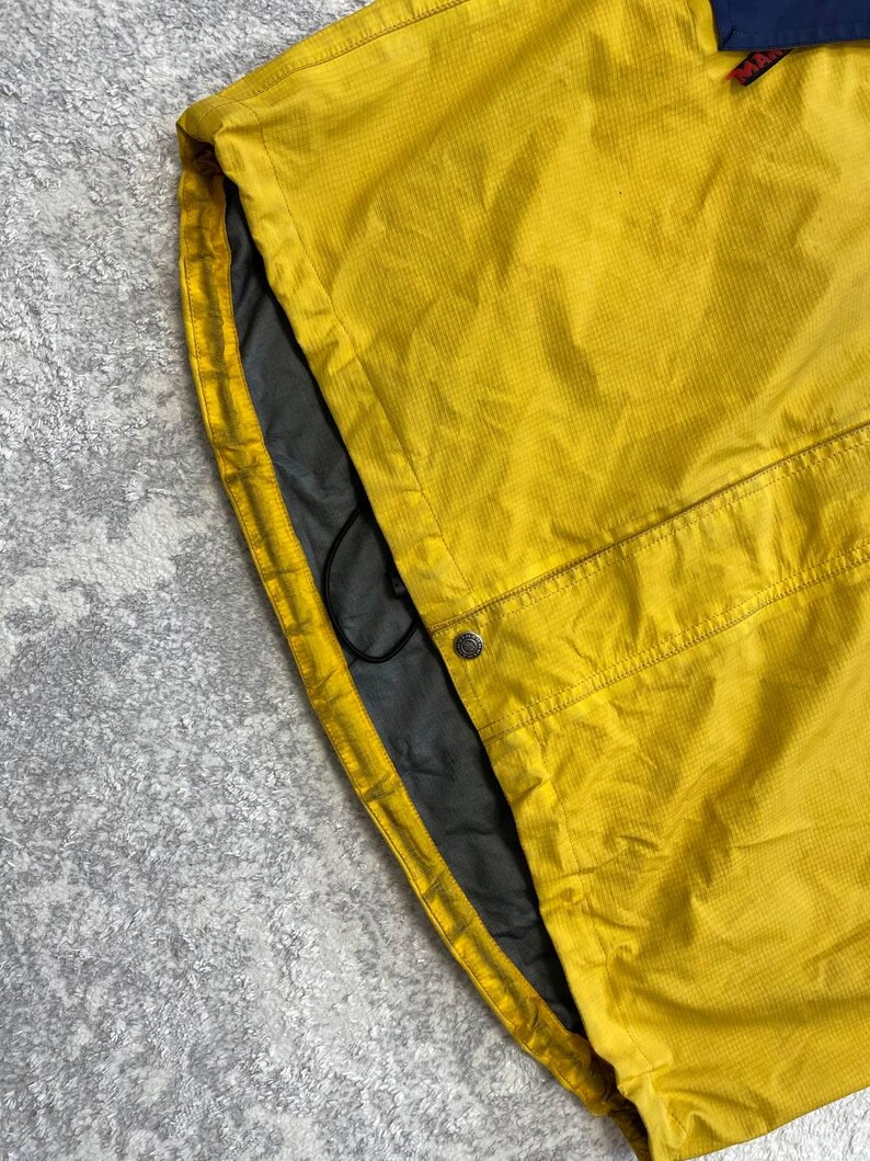May include: A yellow jacket with a grid-like texture, featuring a dark blue panel at the top, a grey interior lining, a black drawstring cord, and a single silver-toned metal snap button closure near the bottom hem area.