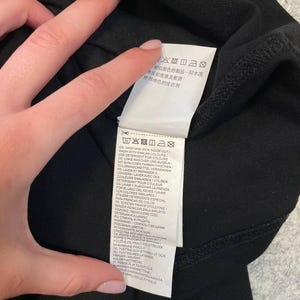 May include: Black garment with a white tag featuring care instructions in multiple languages. The tag includes washing and ironing instructions, along with symbols. The garment is held by a person.