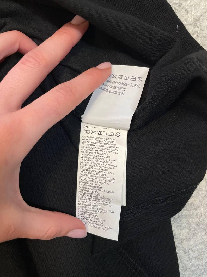 May include: Black garment with a white tag featuring care instructions in multiple languages. The tag includes washing and ironing instructions, along with symbols. The garment is held by a person.