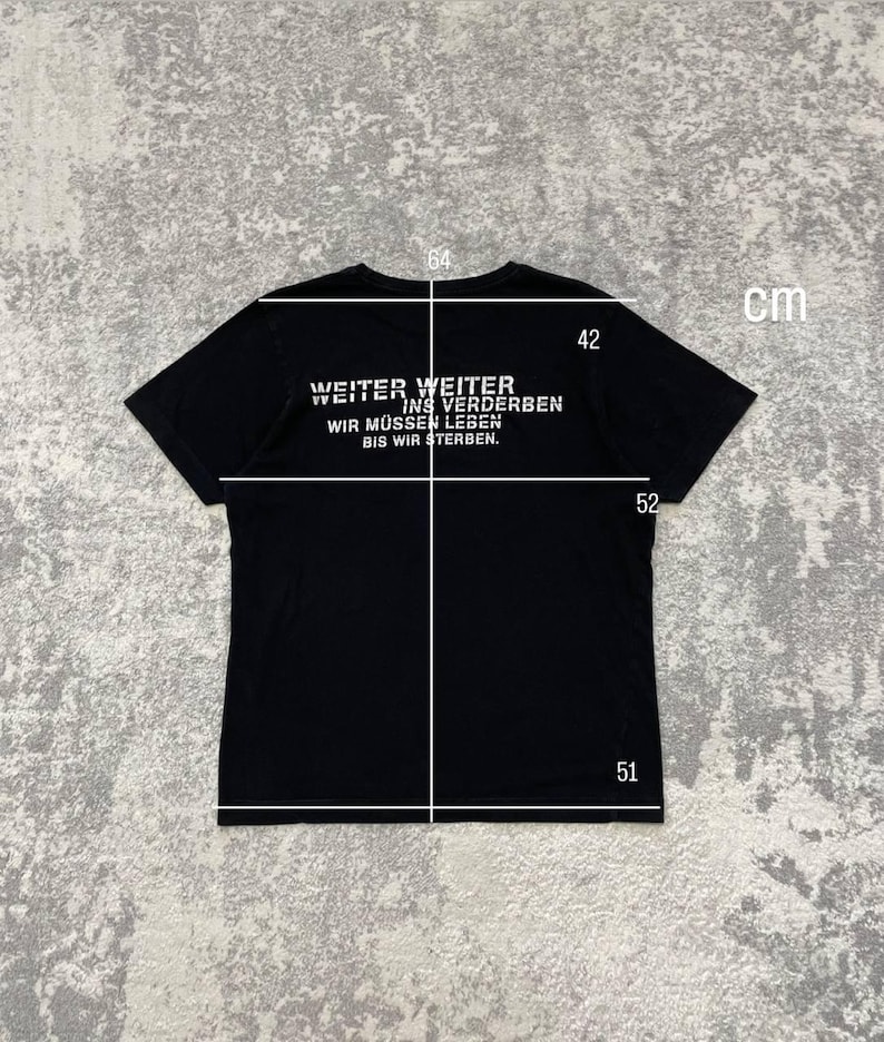 May include: Black t-shirt with white text that reads "WEITER WEITER INS VERDERBEN WIR M&Uuml;SSEN LEBEN BIS WIR STERBEN." The shirt measures 64 cm across the chest, 52 cm in length, 51 cm at the bottom, and 42 cm from the collar to the shoulder. The shirt is displayed on a gray surface.