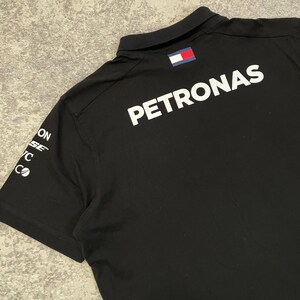 May include: Black polo shirt with the word "PETRONAS" in large white letters on the back. The shirt has a small flag patch on the collar and sponsor logos on the left sleeve. The shirt is made of a smooth, dark fabric.