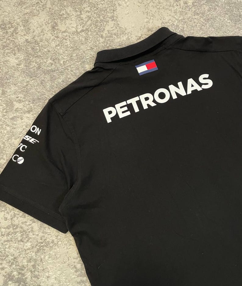 May include: Black polo shirt with the word "PETRONAS" in large white letters on the back. The shirt has a small flag patch on the collar and sponsor logos on the left sleeve. The shirt is made of a smooth, dark fabric.