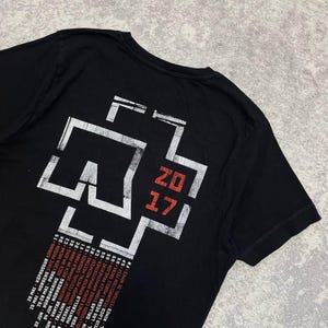 K&ouml;nnte beinhalten: Ein schwarzes T-Shirt mit kurzen &Auml;rmeln und einem gro&szlig;en wei&szlig;-roten Aufdruck auf der R&uuml;ckseite. Das Design zeigt ein stilisiertes Kreuz-Logo, den Text 2017 in Rot sowie eine Liste von Tourdaten und Orten in wei&szlig;er und roter Schrift.