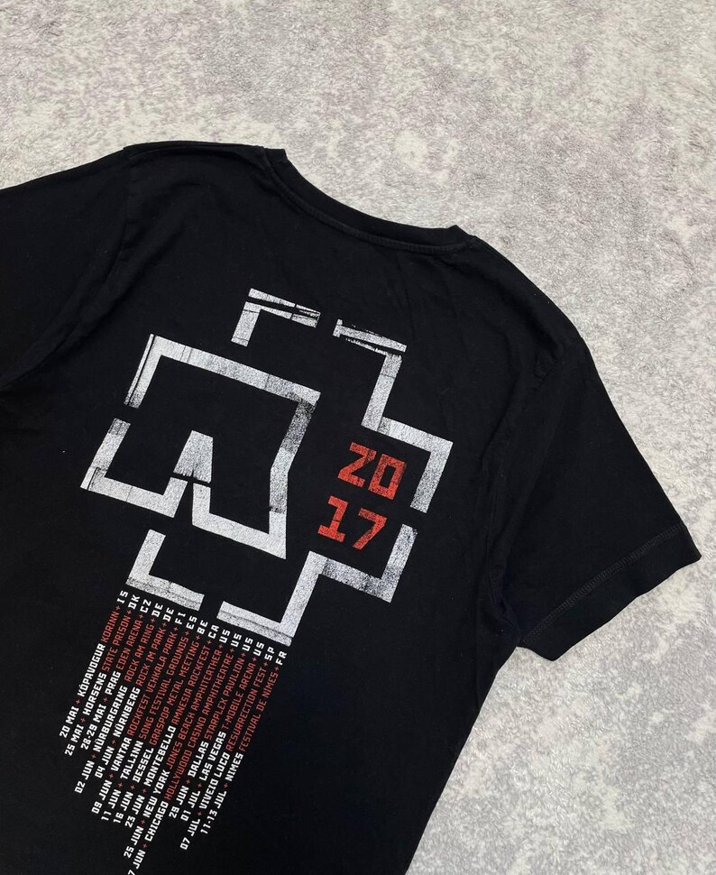 K&ouml;nnte beinhalten: Ein schwarzes T-Shirt mit kurzen &Auml;rmeln und einem gro&szlig;en wei&szlig;-roten Aufdruck auf der R&uuml;ckseite. Das Design zeigt ein stilisiertes Kreuz-Logo, den Text 2017 in Rot sowie eine Liste von Tourdaten und Orten in wei&szlig;er und roter Schrift.