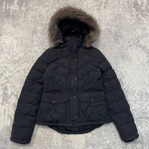 Peut inclure: Une doudoune noire avec un motif matelassé en chevron, une capuche bordée de fausse fourrure brune, une fermeture éclair frontale avec une patte à boutons-pression et deux grandes poches à rabat avec boutons-pression sur le bas du vêtement.