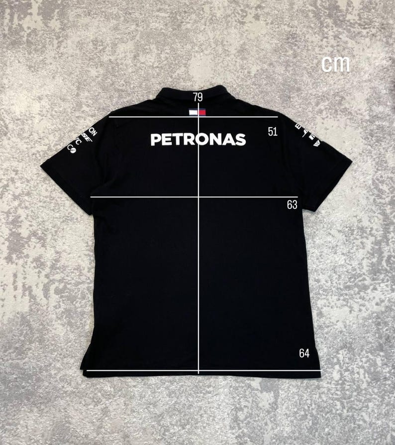 May include: Black polo shirt with the word "PETRONAS" in white across the back. The shirt has short sleeves and a collar. Measurements are shown in centimeters: 79 cm across the shoulders, 51 cm from shoulder to sleeve, 63 cm from shoulder to hem, and 64 cm across the bottom.