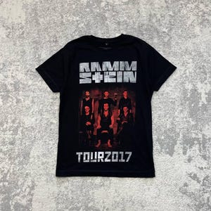 K&ouml;nnte beinhalten: Ein schwarzes Kurzarm-T-Shirt mit dem Bandnamen RAMMSTEIN in einer abgenutzten silbernen Schriftart oben. Unter dem Text befindet sich ein dunkles, stimmungsvolles Foto der sechs Bandmitglieder. Die Worte TOUR 2017 sind unten in Silber gedruckt.