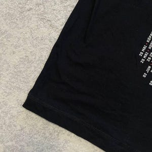 K&ouml;nnte beinhalten: Ein schwarzes T-Shirt mit wei&szlig;em Textaufdruck an der Seite. Der sichtbare Text enth&auml;lt Daten wie 20 MAI + K&Ouml;P, 25 MAI + H&Ouml; und 02 JUN. Das Kleidungsst&uuml;ck hat einen einfachen ges&auml;umten Rand und besteht aus glattem, einfarbig schwarzem Stoff.