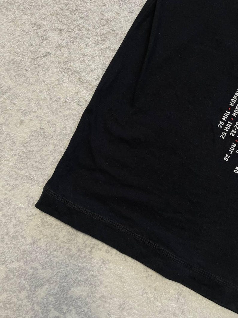 K&ouml;nnte beinhalten: Ein schwarzes T-Shirt mit wei&szlig;em Textaufdruck an der Seite. Der sichtbare Text enth&auml;lt Daten wie 20 MAI + K&Ouml;P, 25 MAI + H&Ouml; und 02 JUN. Das Kleidungsst&uuml;ck hat einen einfachen ges&auml;umten Rand und besteht aus glattem, einfarbig schwarzem Stoff.