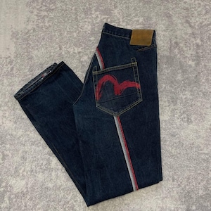 Puede incluir: Vaqueros de mezclilla azul oscuro con una raya roja y plateada en el lateral. El bolsillo trasero presenta un diseño bordado en rojo. Se ve un parche de cuero marrón en la parte trasera de los vaqueros.