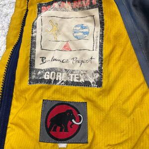May include: The interior of a jacket featuring a bright yellow lining and a dark blue zipper. A rectangular patch displays the text MAMMUT, Balance Project, and GORE-TEX, with a graphic of a figure and a globe. A second patch shows a black mammoth logo.