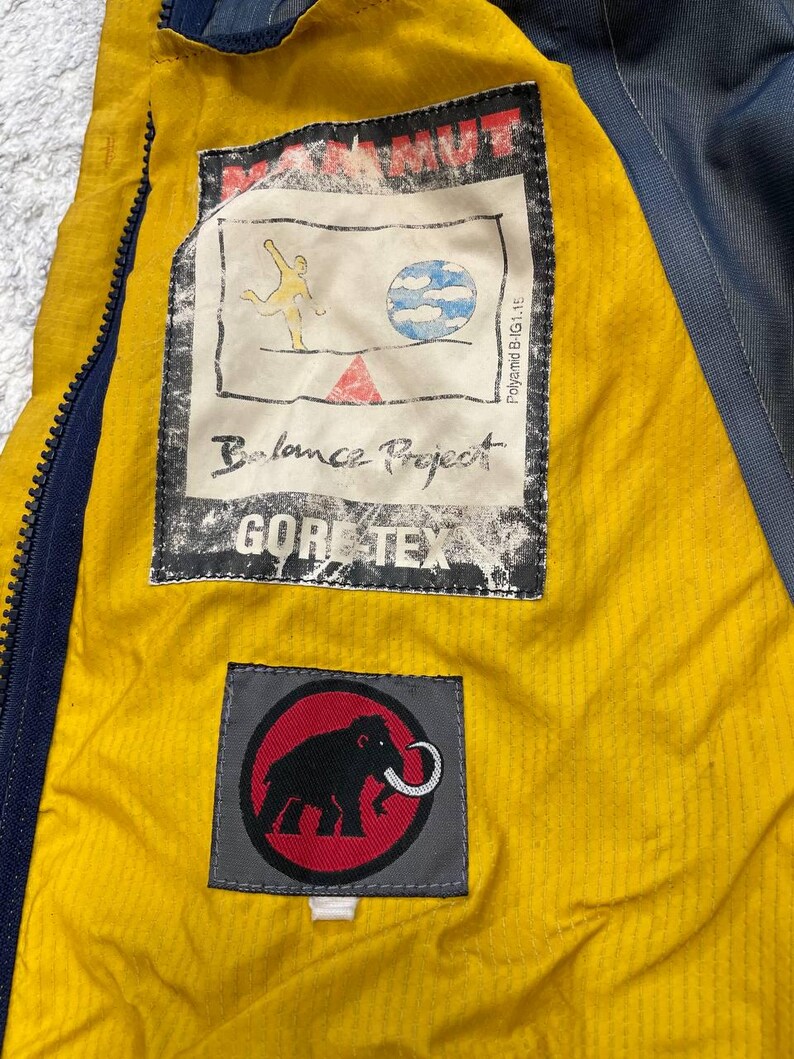 May include: The interior of a jacket featuring a bright yellow lining and a dark blue zipper. A rectangular patch displays the text MAMMUT, Balance Project, and GORE-TEX, with a graphic of a figure and a globe. A second patch shows a black mammoth logo.