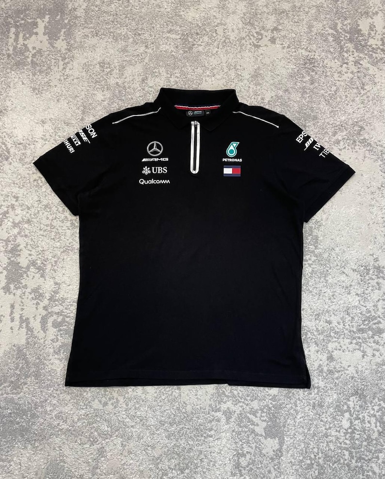 May include: Black polo shirt with a white zipper and collar trim. Features logos for AMG, UBS, Qualcomm, and Petronas. Short sleeves with sponsor logos. A sporty, casual top.