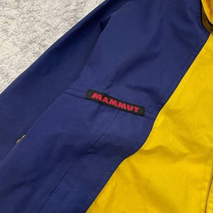 May include: A color-blocked jacket featuring a deep blue sleeve and a bright yellow front panel. A black rectangular patch with the red text MAMMUT is stitched onto the upper sleeve area. The fabric has a subtle grid-like texture throughout.