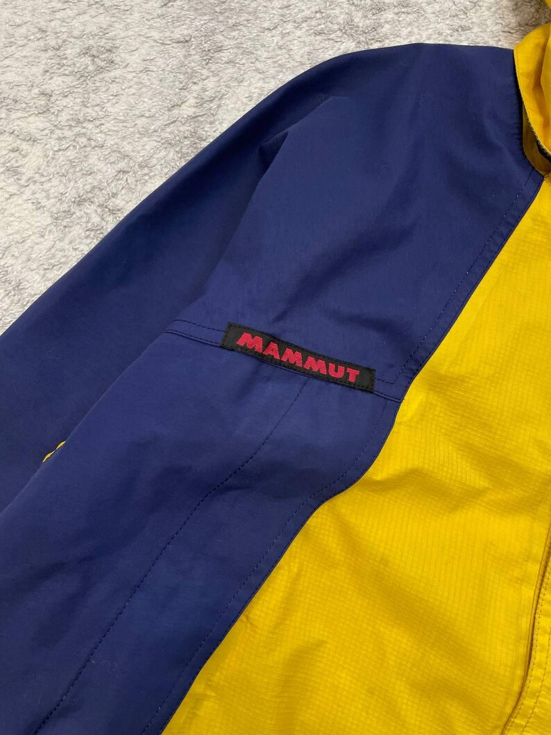 May include: A color-blocked jacket featuring a deep blue sleeve and a bright yellow front panel. A black rectangular patch with the red text MAMMUT is stitched onto the upper sleeve area. The fabric has a subtle grid-like texture throughout.