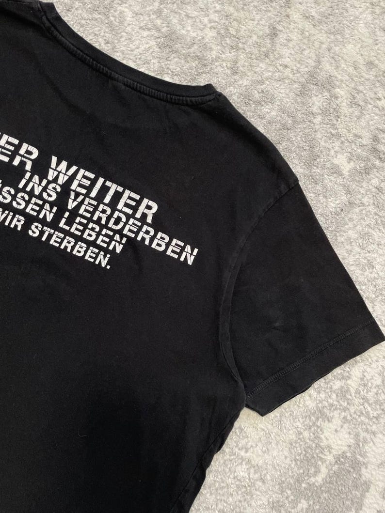 May include: Black t-shirt with white text. The text reads "ER WEITER INS VERDERBEN MUSSEN LEBEN WIR STERBEN." The shirt has a crew neck and short sleeves. The fabric appears to be cotton or a cotton blend.
