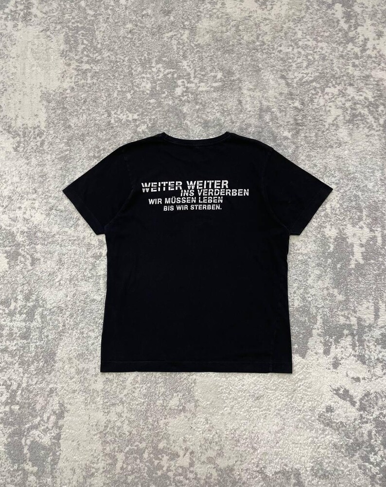 May include: Black t-shirt with white text on the back. The text reads "WEITER WEITER INS VERDERBEN WIR M&Uuml;SSEN LEBEN BIS WIR STERBEN."