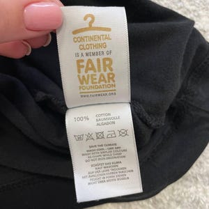 May include: Close-up of a black garment tag with the text "CONTINENTAL CLOTHING IS A MEMBER OF FAIR WEAR FOUNDATION". The tag also includes washing instructions and the text "100% COTTON".
