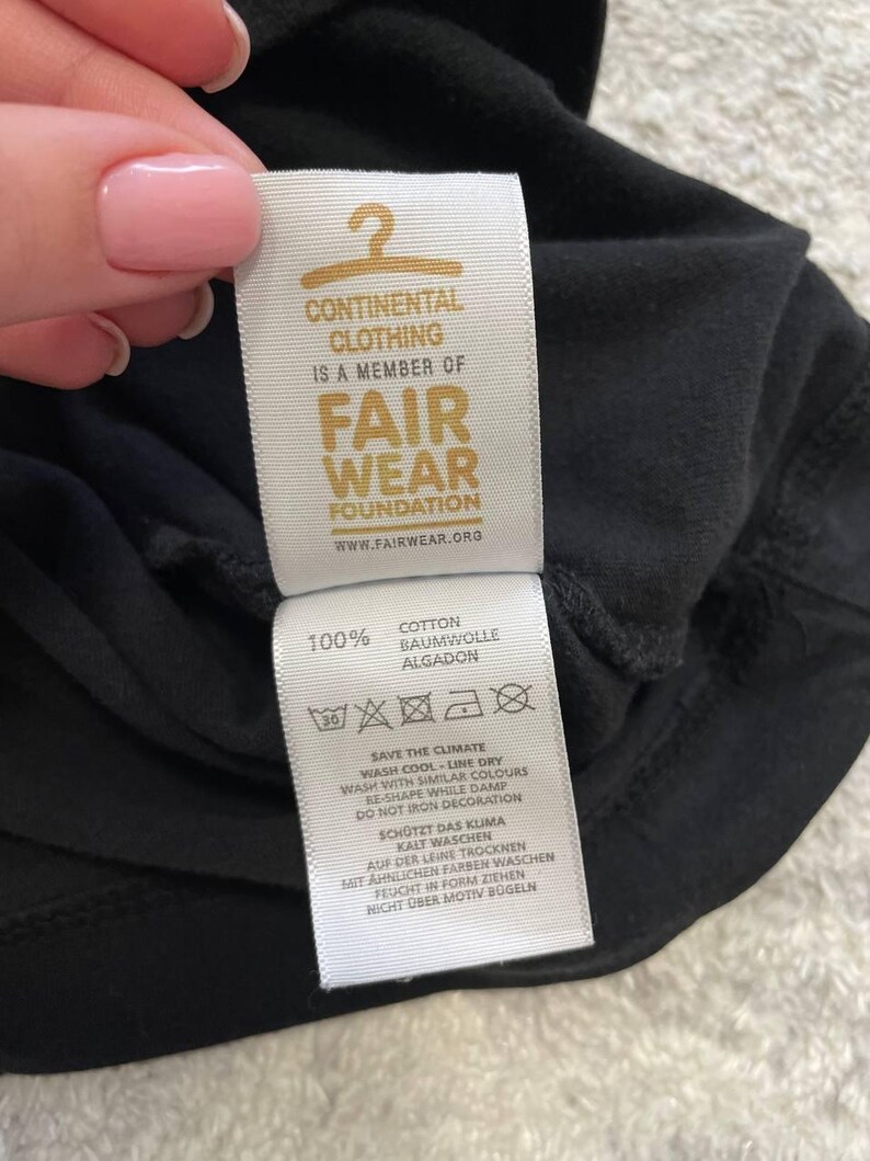 May include: Close-up of a black garment tag with the text "CONTINENTAL CLOTHING IS A MEMBER OF FAIR WEAR FOUNDATION". The tag also includes washing instructions and the text "100% COTTON".