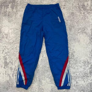 Pode incluir: Calças desportivas azuis com cintura elástica e punhos nos tornozelos. As calças apresentam um logótipo adidas branco na coxa esquerda e riscas diagonais decorativas vermelhas e brancas ao longo das pernas com três riscas verticais.
