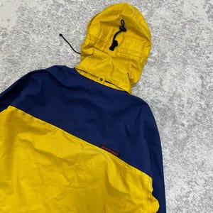 May include: A color-blocked jacket featuring a bright yellow hood with a black drawstring toggle and a yellow lower section contrasted by a dark blue upper back and shoulder area. The word Mammut is printed in small red letters on the blue fabric.