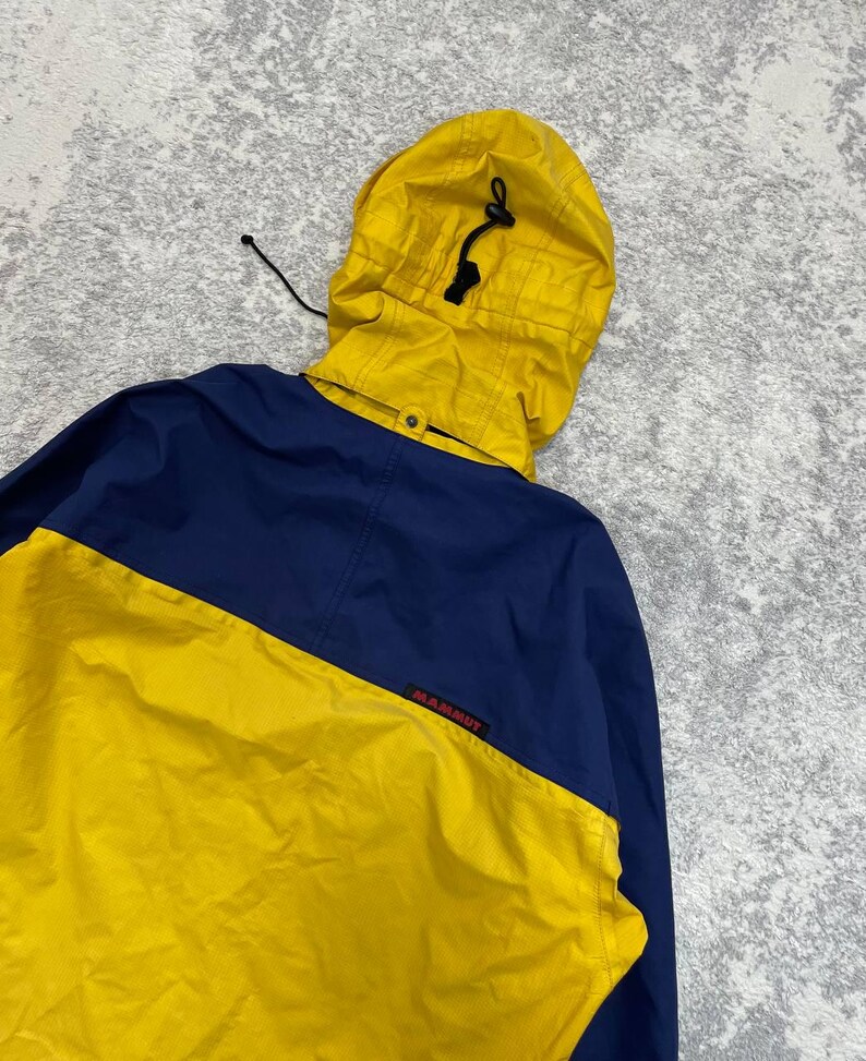 May include: A color-blocked jacket featuring a bright yellow hood with a black drawstring toggle and a yellow lower section contrasted by a dark blue upper back and shoulder area. The word Mammut is printed in small red letters on the blue fabric.